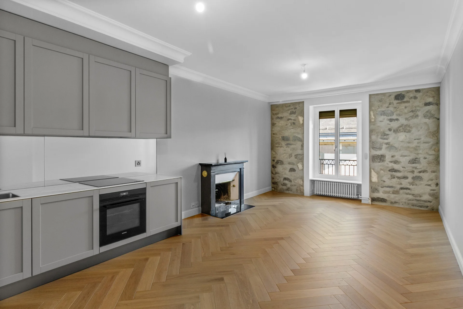 Séjour avec cuisine ouverte après rénovation haut de gamme à Genève, parquet en chêne chevron, cheminée et mur en pierre d’origine